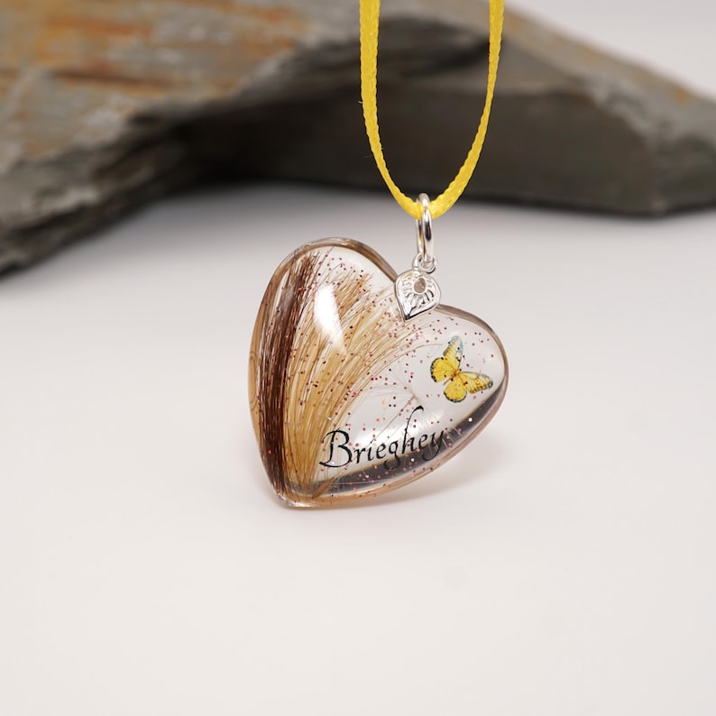 May include: Heart-shaped pendant with a yellow ribbon. The clear resin pendant contains hair and a yellow butterfly design. The name "Briechey" is written on the pendant. The pendant is suspended from a yellow ribbon.
