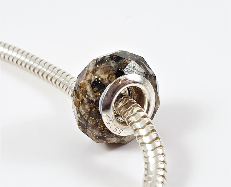 May include: A silver charm bracelet bead with a dark brown and gold speckled glass insert. The bead is faceted and has a round hole for threading onto a bracelet. The bead is marked with the number "S925" on the side.