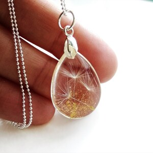 May include: A silver chain necklace with a teardrop-shaped pendant. The pendant is filled with a clear resin and contains a dried dandelion with gold glitter.