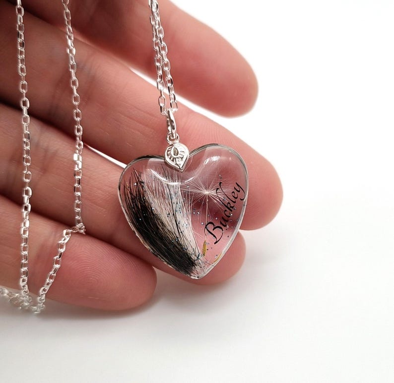 May include: Silver heart-shaped pendant necklace with a clear resin center. The pendant contains black and white hair and the name "Buckley" in cursive. The necklace is on a silver chain.