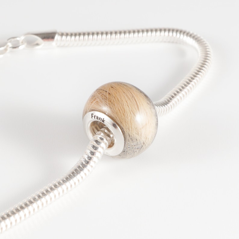 May include: A silver chain bracelet with a round bead filled with light brown hair. The bead is engraved with the name "Frank".