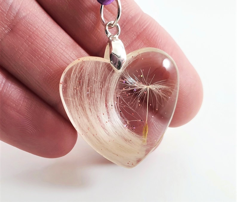 May include: Heart-shaped resin pendant with a dandelion seed head and light-colored hair inclusions. The clear resin is accented with pink glitter. The pendant hangs from a purple cord and silver-colored bail.
