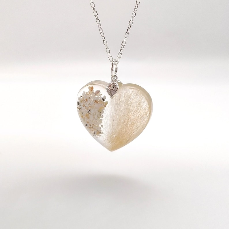 May include: A silver chain necklace with a heart-shaped pendant. The pendant is made of clear resin and contains a mixture of sand and hair. The pendant is attached to the chain with a small silver charm.