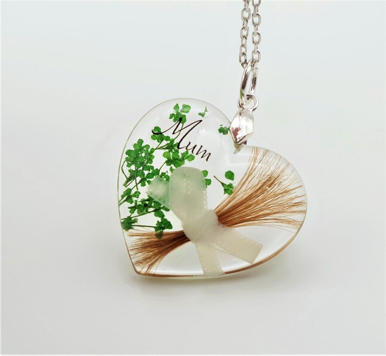 May include: A heart-shaped pendant necklace with a clear resin design. The pendant contains brown hair, green foliage, and a white ribbon. The word "Mum" is written in black script. The necklace has a silver chain.