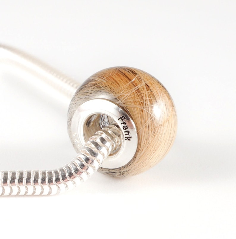 May include: A silver charm bracelet with a round bead made of clear resin filled with light brown hair. The bead is engraved with the name "Frank".