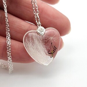 May include: A silver necklace with a heart-shaped pendant. The clear pendant contains white fluffy material and the word "Salt" in black script. The necklace is held in a hand.
