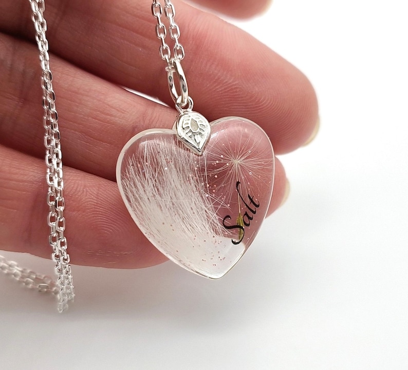 May include: A silver necklace with a heart-shaped pendant. The clear pendant contains white fluffy material and the word "Salt" in black script. The necklace is held in a hand.