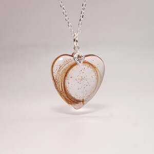 May include: A heart-shaped pendant necklace with a clear resin heart. The heart contains strands of brown hair and red glitter. The pendant hangs from a silver chain. The pendant has a silver bail.
