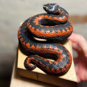 Puede incluir: Una figura de serpiente enroscada con un patrón texturizado en negro y naranja quemado en todo el cuerpo. La escultura tiene una cabeza detallada con ojos visibles y descansa sobre una base de bloque de madera clara con escamas realistas.