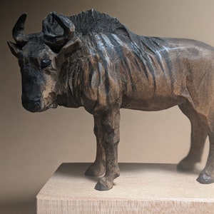 Puede incluir: Una talla de madera de un ñu, un gran antílope africano, de pie sobre una base de madera. La talla es detallada y realista, con un esquema de color marrón oscuro y negro.