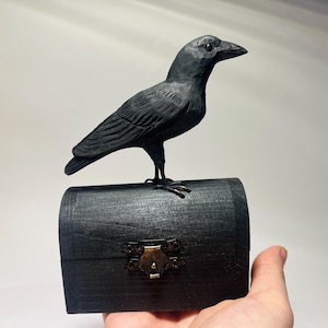 Puede incluir: Un cuervo negro tallado posado sobre un pequeño cofre de madera negro. El cofre tiene un cierre de metal. El cuervo está detallado con plumas talladas y un pico afilado. La estética general es gótica y misteriosa.