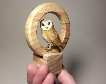 May include: A wooden sculpture of a barn owl perched inside a wooden ring. The owl is painted with realistic details, including brown and white feathers and black eyes.