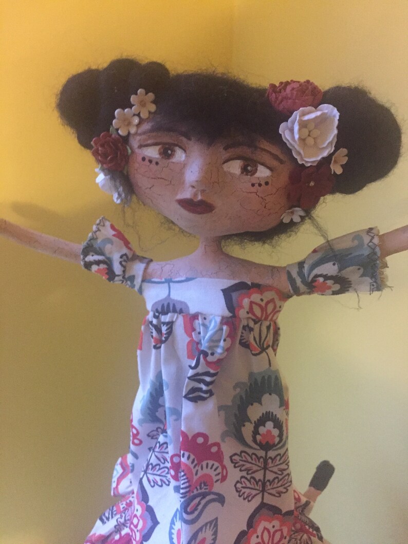 May include: A handmade doll with black hair, brown eyes, and a floral dress. The doll is wearing a flower crown with red, white, and brown flowers.