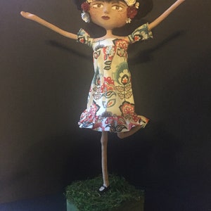 May include: A handmade doll with a floral dress and flowers in her hair. The doll is standing on a small wooden base covered in green moss.