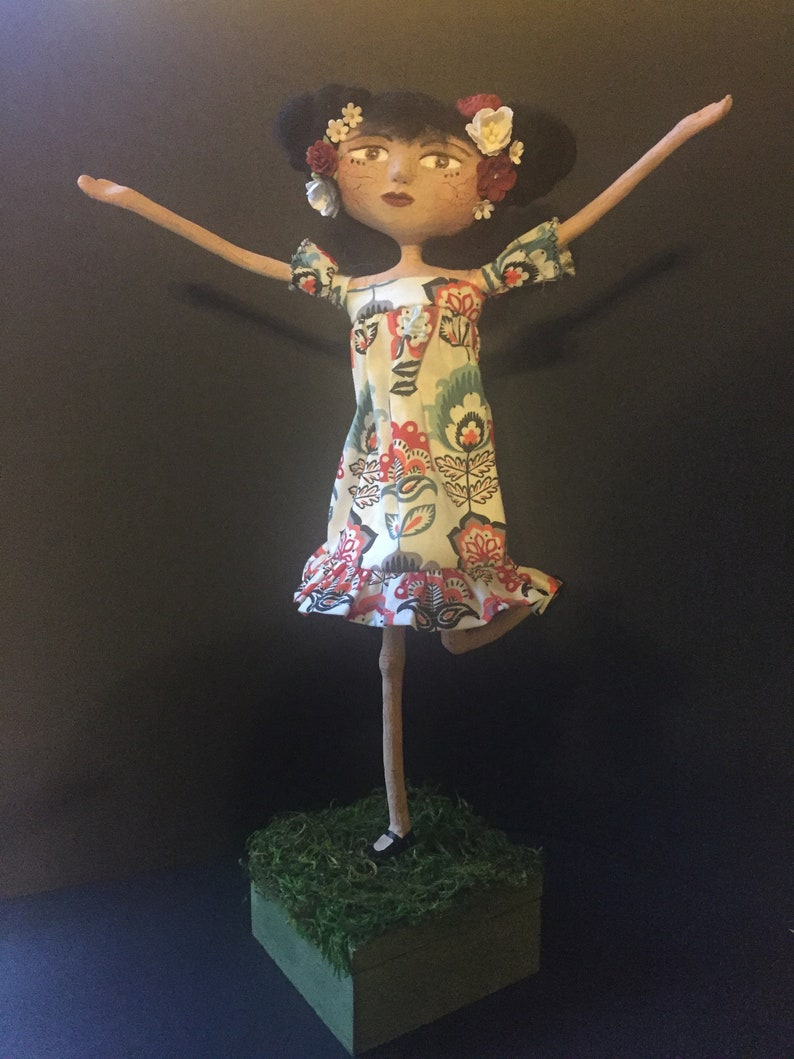 May include: A handmade doll with a floral dress and flowers in her hair. The doll is standing on a small wooden base covered in green moss.