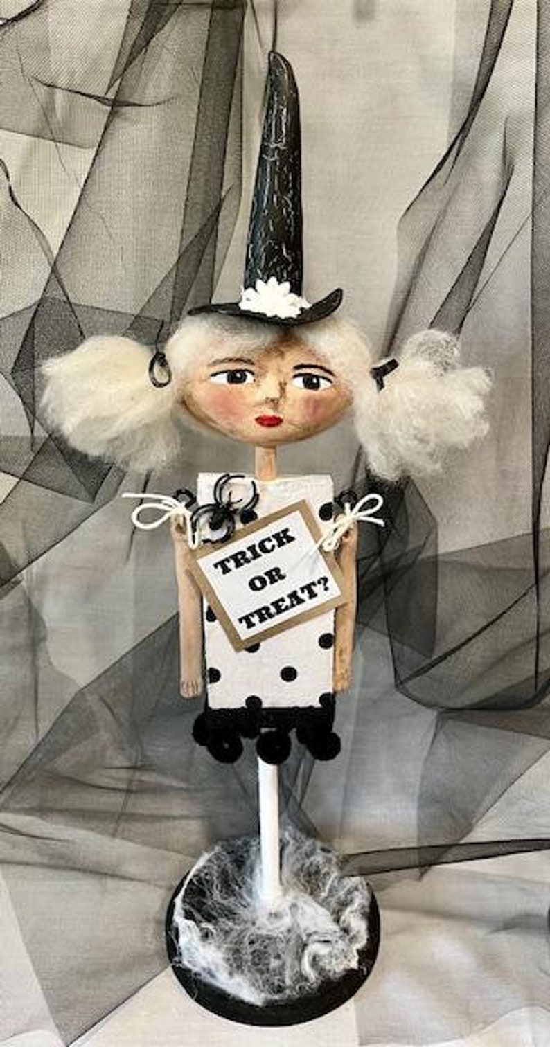 May include: A white witch doll with black polka dot dress and a black witch hat with white flowers. The doll is holding a sign that says "Trick or Treat?" and is standing on a black base with white spiderwebs.