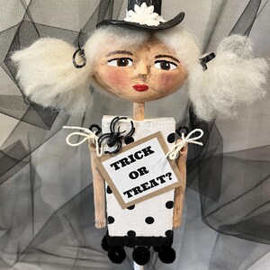 May include: A white and black polka dot doll with white hair, a black hat with a white flower, and a sign that says "Trick or Treat?"