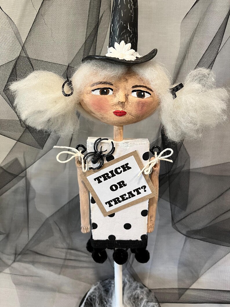 May include: A white and black polka dot doll with white hair, a black hat with a white flower, and a sign that says "Trick or Treat?"