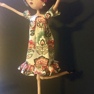 May include: A handmade doll with long, slender limbs, wearing a white dress with a floral pattern in shades of red, green, and black. The doll has black hair and is adorned with red, white, and pink flowers.