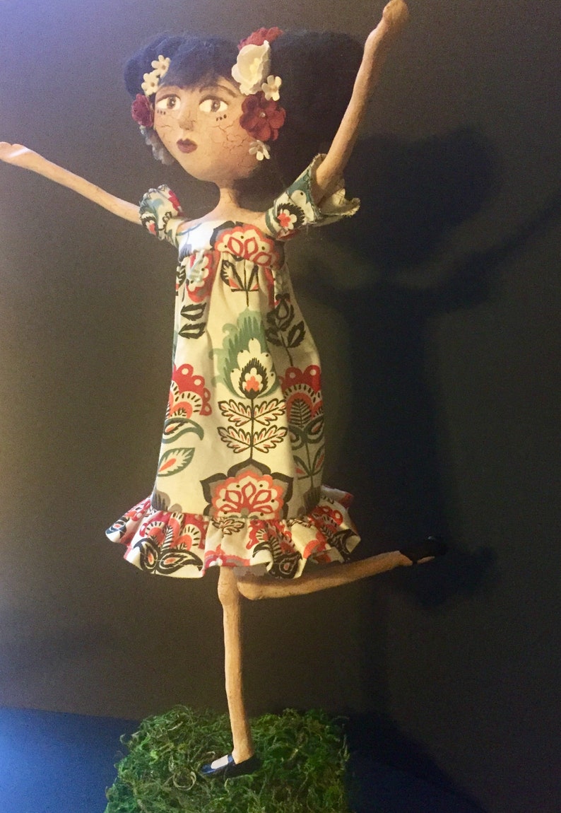 May include: A handmade doll with long, slender limbs, wearing a white dress with a floral pattern in shades of red, green, and black. The doll has black hair and is adorned with red, white, and pink flowers.