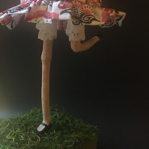 May include: A handmade doll with a white dress with red and black floral print, white lace trim, and a brown stick leg. The doll is standing on a bed of green moss.