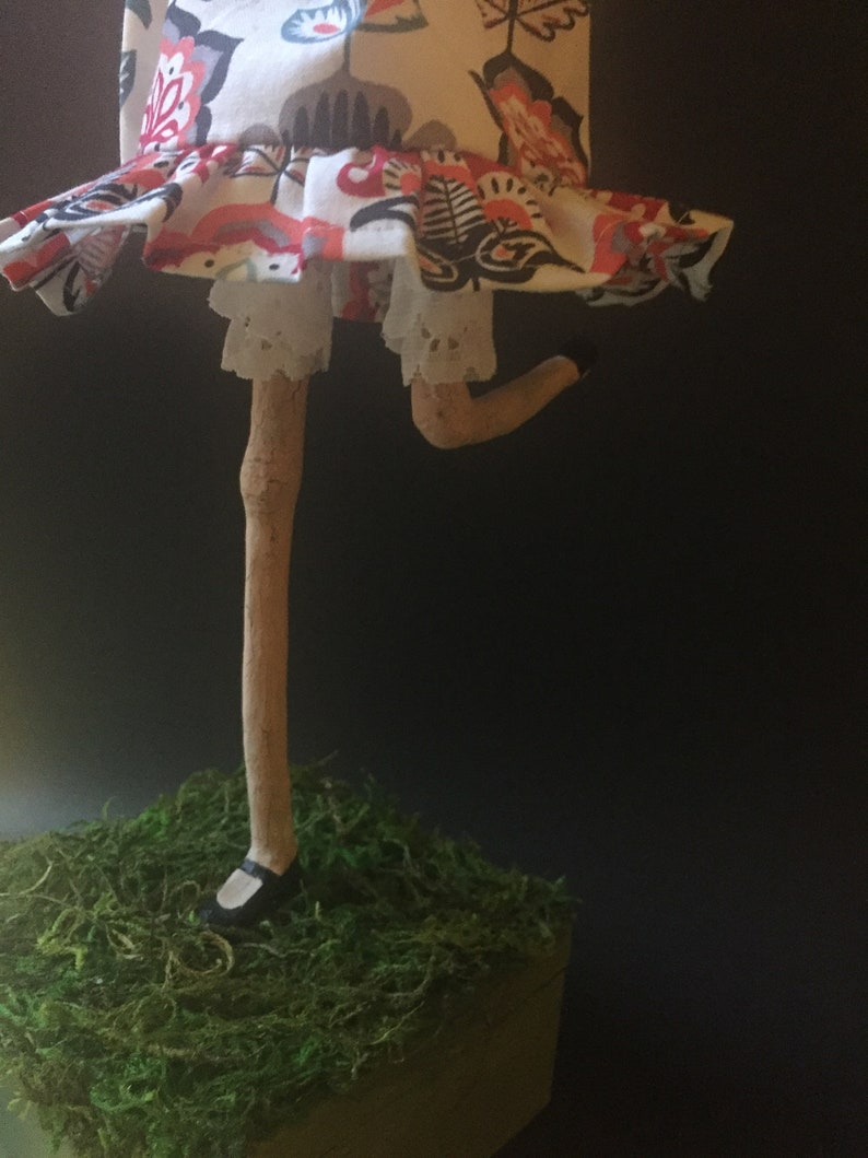 May include: A handmade doll with a white dress with red and black floral print, white lace trim, and a brown stick leg. The doll is standing on a bed of green moss.