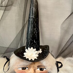 May include: A white haired doll with a black and white witch hat with a white flower on the brim. The doll has a painted face with brown eyes and red lips.