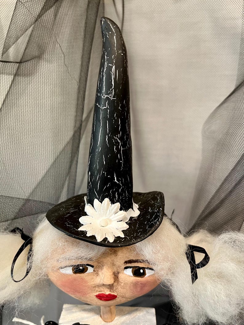 May include: A white haired doll with a black and white witch hat with a white flower on the brim. The doll has a painted face with brown eyes and red lips.