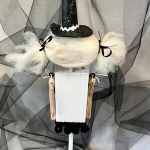 May include: A white witch doll with black hair and a black witch hat. The doll is standing on a black base with white spiderwebs.