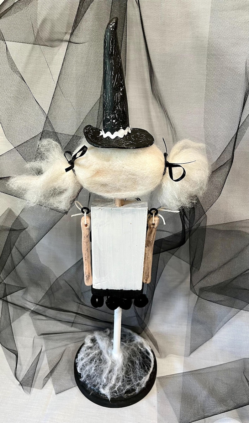 May include: A white witch doll with black hair and a black witch hat. The doll is standing on a black base with white spiderwebs.