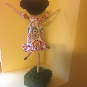 May include: A handmade doll with black hair, wearing a white dress with red and blue floral patterns, standing on a green base.