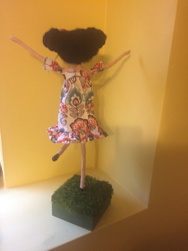 May include: A handmade doll with black hair, wearing a white dress with red and blue floral patterns, standing on a green base.