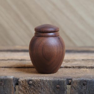 May include: A small, round wooden jar with a lid. The jar is made of dark brown wood and has a smooth, polished finish.