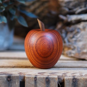 May include: A wooden apple sculpture with a reddish-brown finish. The apple has a smooth, rounded surface with visible wood grain.
