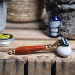 May include: A wooden handled safety razor with a gold colored head and a blue and green razor blade. The razor is resting on a gray rock.