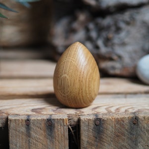 May include: A wooden egg-shaped ornament with a light brown colour and a wood grain pattern. The ornament is sitting on a wooden surface.