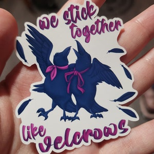 May include: A white sticker with two blue crows with pink ribbons around their necks. The crows are facing each other and have their wings spread. The text "we stick together like velcro" is written in pink on the sticker.
