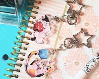 May include: Two clear acrylic keychains featuring a cat design. One keychain has a cat holding a red 20-sided die with white numbers. The other keychain has a cat holding a blue 20-sided die with white numbers. Both keychains have a silver star-shaped clasp.