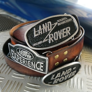 Może przedstawiać: Brązowy skórzany pasek z trzema oddzielnymi metalowymi klamrami. Klamry prezentują logo Land Rover, napis Land Rover Solihull Warwickshire England oraz napis Land Rover Experience wykonany srebrnymi literami na czarnym tle.