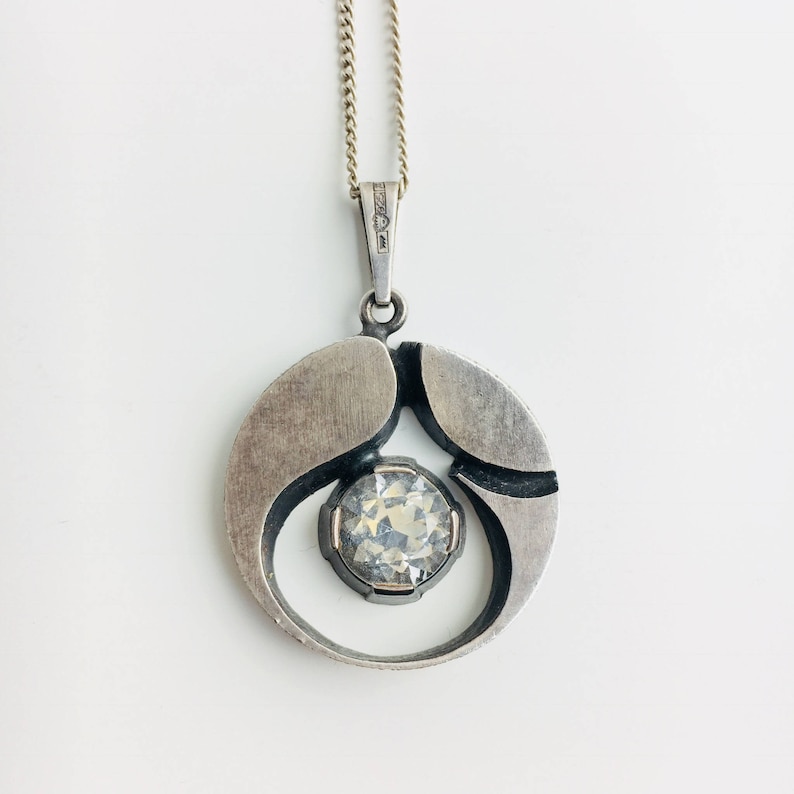 May include: A silver pendant necklace with a round, abstract design featuring a clear crystal in the center. The pendant is suspended from a silver chain.