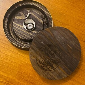 May include: A wooden box with a dark brown finish and a circular design engraved on the lid. The box is open and a silver necklace with a crystal pendant is inside.