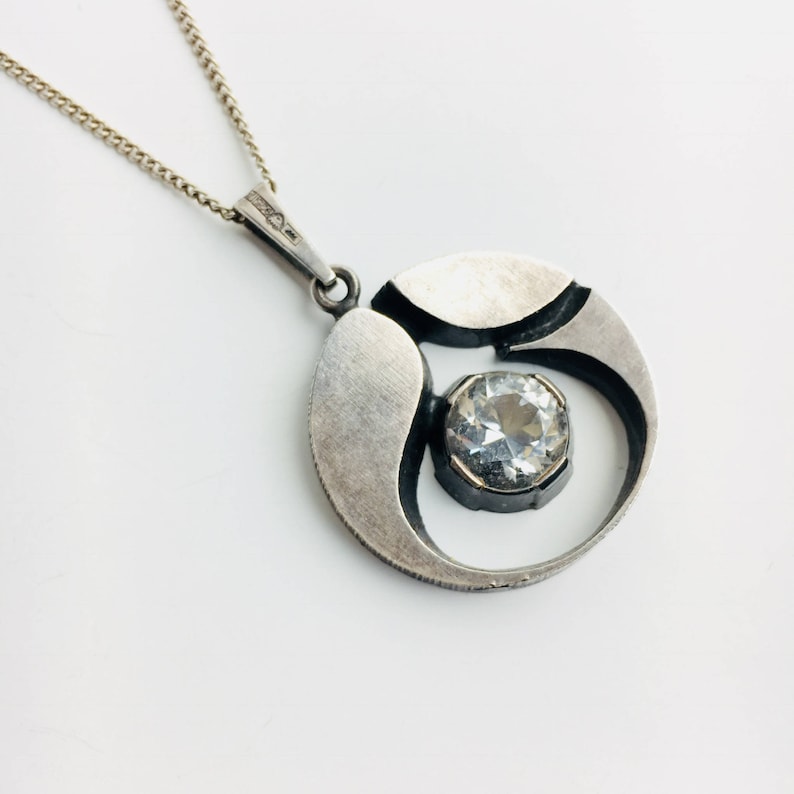 May include: A silver pendant necklace with a round, abstract design featuring a clear crystal stone in the center.