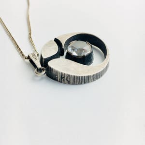 May include: A silver necklace with a circular pendant featuring a clear crystal stone set in a silver frame. The pendant has a textured surface and the words "KL" are visible on the back.