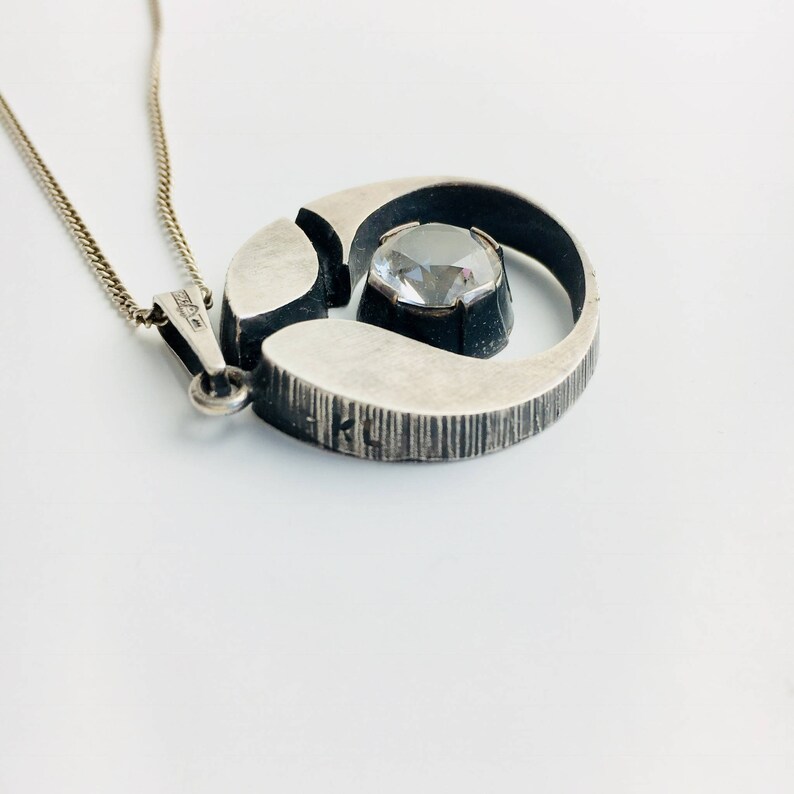 May include: A silver necklace with a circular pendant featuring a clear crystal stone set in a silver frame. The pendant has a textured surface and the words "KL" are visible on the back.