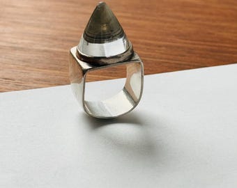 May include: A silver ring with a clear, pointed crystal set in a square bezel.