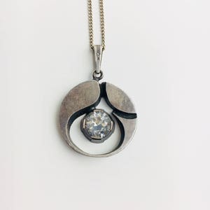 May include: A silver pendant necklace with a round, abstract design featuring a clear crystal in the center.