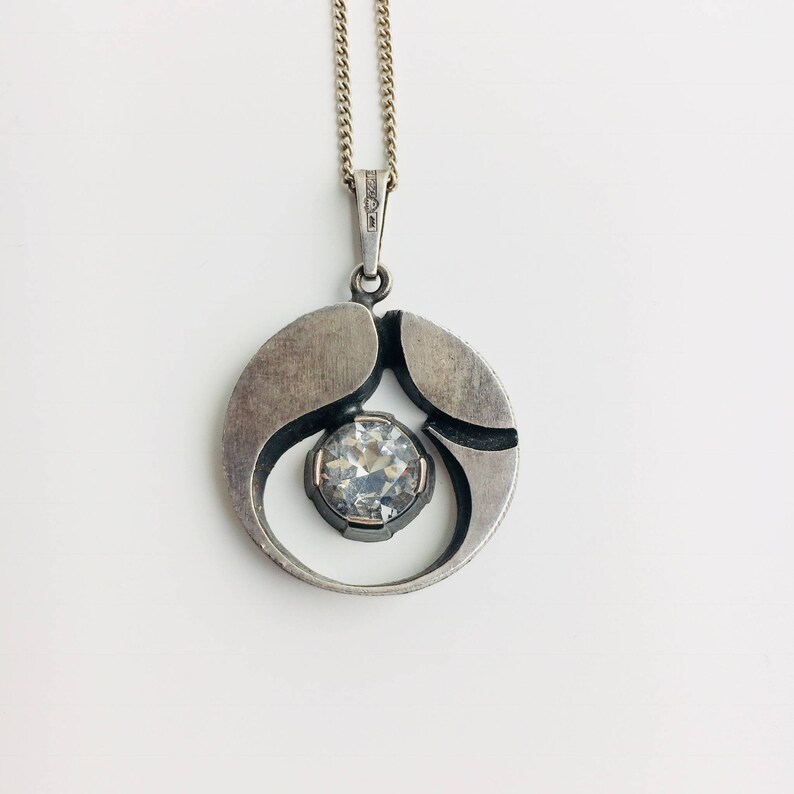 May include: A silver pendant necklace with a round, abstract design featuring a clear crystal in the center.