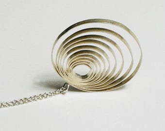 Pode incluir: Um colar com pingente prateado com um design em espiral. O pingente é composto por círculos concêntricos, criando um efeito tridimensional. O colar tem uma corrente delicada e o pingente está centrado. O fundo é branco liso.