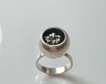 May include: A silver ring with a round, hollow top that contains a cluster of small, silver beads.