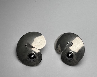 May include: A pair of silver-toned metal earrings featuring a smooth, spiraling, shell-like shape. Each earring is accented with a single, round, polished black stone set securely in the center of the spiral, creating a reflective and metallic finish.